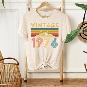 May include: Cream-colored t-shirt with a retro design. The design features the word "VINTAGE" in gold, a mountain scene, and the number "1976" in colorful stripes. The shirt is hanging on a wooden rack.