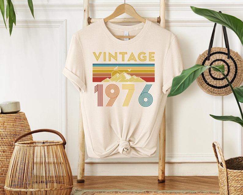 May include: Cream-colored t-shirt with a retro design. The design features the word "VINTAGE" in gold, a mountain scene, and the number "1976" in colorful stripes. The shirt is hanging on a wooden rack.