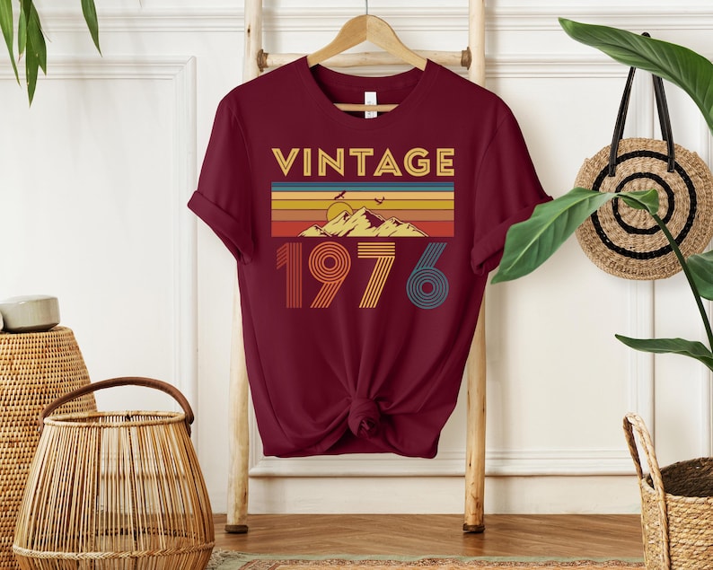 May include: Burgundy t-shirt with a retro design. The graphic features the word "VINTAGE" above a mountain scene with a sunset and the year "1976" in a colorful, striped font. The shirt is hanging on a wooden hanger.