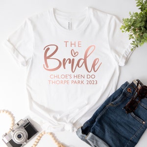 May include: White t-shirt with the text "The Bride" in rose gold lettering, with a small heart above the word "Bride". Below the text is "Chloe's Hen Do Thorpe Park 2023" in rose gold lettering.