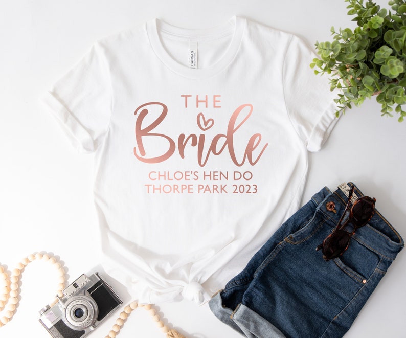 May include: White t-shirt with the text "The Bride" in rose gold lettering, with a small heart above the word "Bride". Below the text is "Chloe's Hen Do Thorpe Park 2023" in rose gold lettering.
