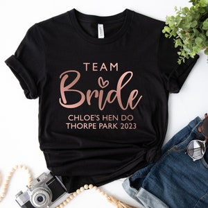 May include: Black t-shirt with rose gold text that reads "Team Bride Chloe's Hen Do Thorpe Park 2023".
