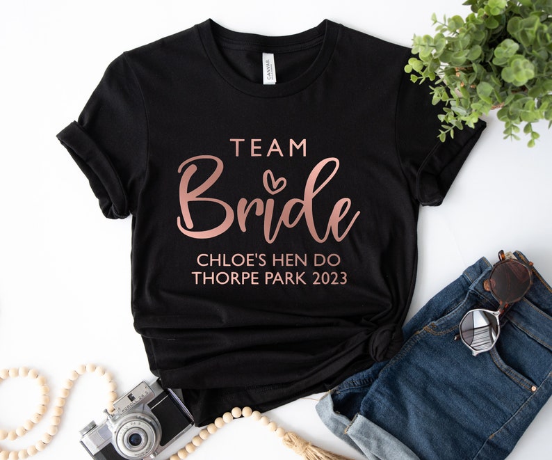 May include: Black t-shirt with rose gold text that reads "Team Bride Chloe's Hen Do Thorpe Park 2023".