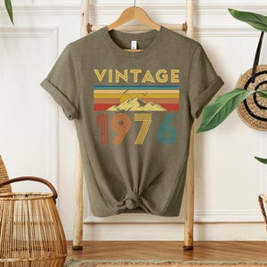 May include: Olive green t-shirt with the word "VINTAGE" in gold letters above a colorful mountain and sunset design. The number "1976" is printed below in orange, yellow, and teal.