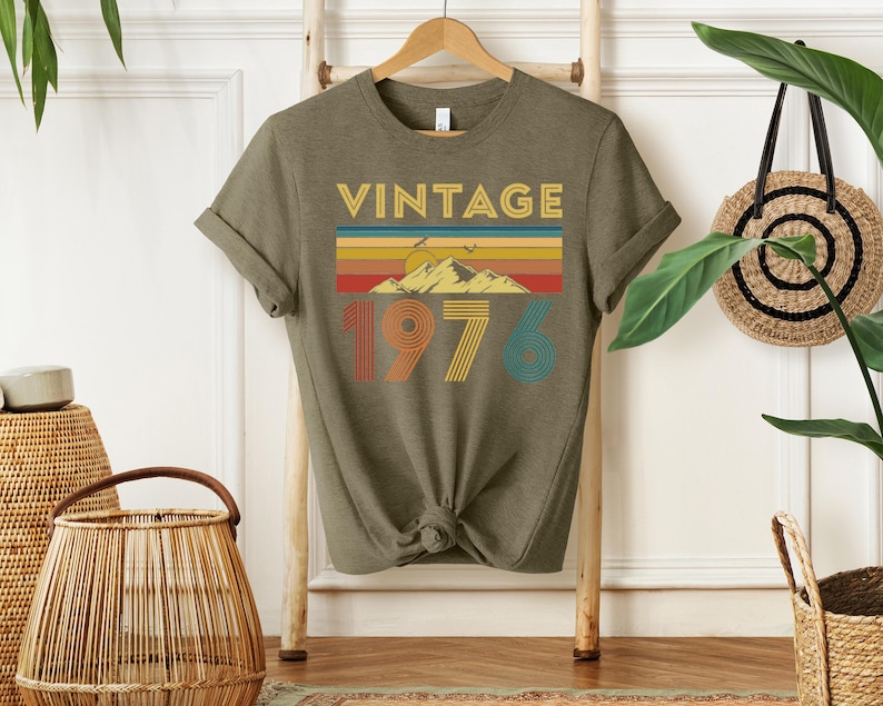 May include: Olive green t-shirt with the word "VINTAGE" in gold letters above a colorful mountain and sunset design. The number "1976" is printed below in orange, yellow, and teal.