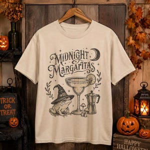 May include: A beige short-sleeve t-shirt featuring a black graphic print with the text Midnight Margaritas. The design includes a frog wearing a witch hat, a margarita glass, a coffee percolator, a crescent moon, stars, and small mushrooms.