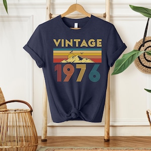 May include: Navy blue t-shirt with a retro design. The design features the word "VINTAGE" in gold, a mountain scene, and the year "1976" in red, orange, yellow, and teal. The shirt is knotted at the waist.