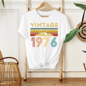 May include: White t-shirt with the word "VINTAGE" in gold letters above a retro design of mountains and stripes in yellow, orange, and blue. The number "1976" is printed below in red, yellow, and teal.