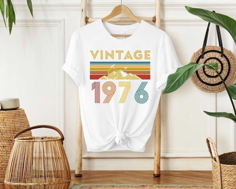 May include: White t-shirt with the word "VINTAGE" in gold letters above a retro design of mountains and stripes in yellow, orange, and blue. The number "1976" is printed below in red, yellow, and teal.