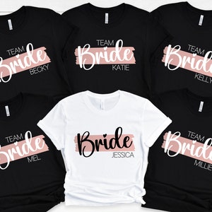 May include: Five black t-shirts with a white graphic of the word "Bride" with a pink brushstroke behind it. Each shirt has the word "Team" and a name below the graphic. The names are Becky, Katie, Kelly, Mel, and Millie. One white t-shirt has the same graphic with the name Jessica.