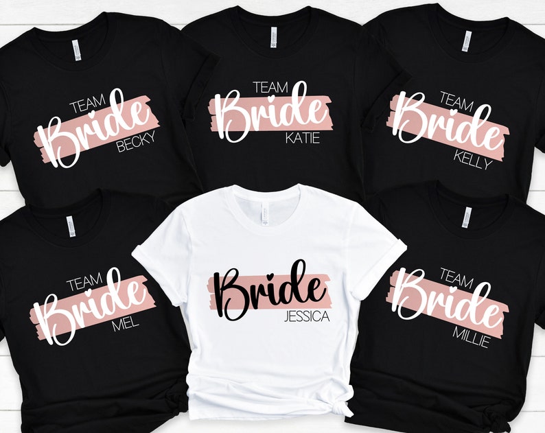 May include: Five black t-shirts with a white graphic of the word "Bride" with a pink brushstroke behind it. Each shirt has the word "Team" and a name below the graphic. The names are Becky, Katie, Kelly, Mel, and Millie. One white t-shirt has the same graphic with the name Jessica.