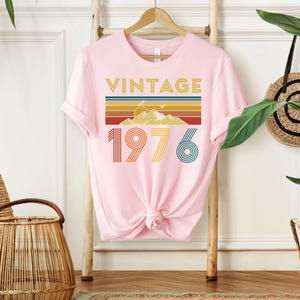 May include: A light pink t-shirt with the word "VINTAGE" in gold letters above a retro design of a mountain and stripes in various colors. Below the design is the number "1976" in a retro font.