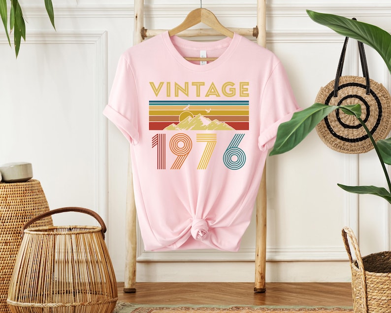 May include: A light pink t-shirt with the word "VINTAGE" in gold letters above a retro design of a mountain and stripes in various colors. Below the design is the number "1976" in a retro font.
