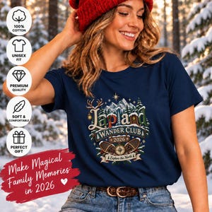 May include: A navy blue short-sleeve t-shirt featuring a graphic print with the text Lapland Wander Club 2026 Explore the North. The design includes snowy mountains, pine trees, a lantern, a small cabin, and snowshoes with a compass centre.