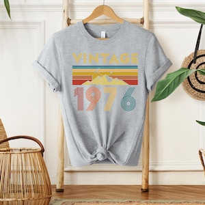 May include: A heather gray t-shirt with a retro design. The design features the word "VINTAGE" above a horizontal stripe pattern in yellow, orange, red, and blue. Below the stripes are the numbers "1976" in orange and teal.