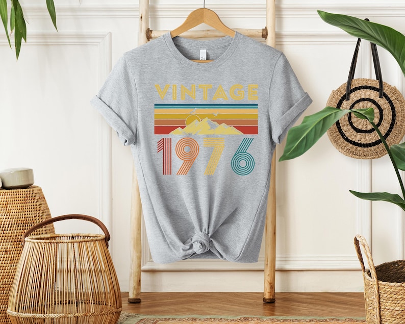 May include: A heather gray t-shirt with a retro design. The design features the word "VINTAGE" above a horizontal stripe pattern in yellow, orange, red, and blue. Below the stripes are the numbers "1976" in orange and teal.