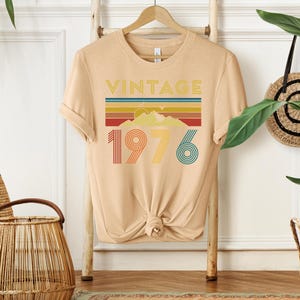 May include: A tan t-shirt with the word "VINTAGE" above a colorful mountain and stripe design. Below the design is the number "1976" in red, yellow, and teal. The shirt is hanging on a wooden hanger.