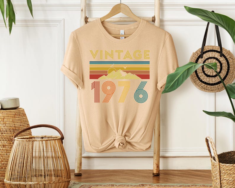 May include: A tan t-shirt with the word "VINTAGE" above a colorful mountain and stripe design. Below the design is the number "1976" in red, yellow, and teal. The shirt is hanging on a wooden hanger.
