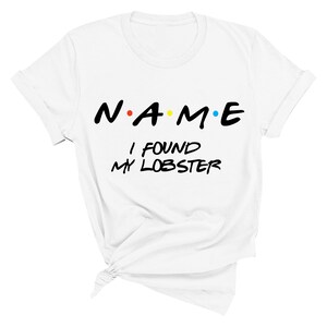 May include: White t-shirt with black text that reads "N.A.M.E. I FOUND MY LOBSTER". The letters in "NAME" are in a stylized font and have colored dots between them.