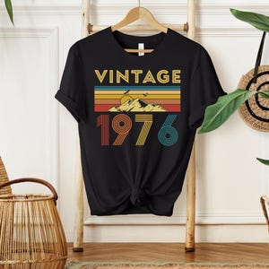 May include: Black t-shirt with the word "VINTAGE" in gold letters above a retro mountain and sunset design. Below the design is the number "1976" in orange, yellow, and teal. The shirt is hanging on a wooden hanger.