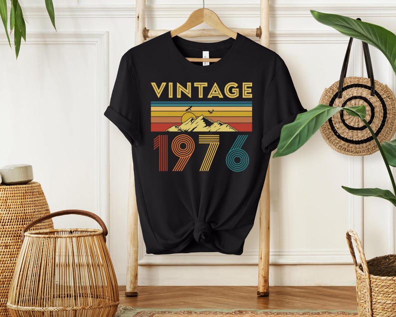 May include: Black t-shirt with the word "VINTAGE" in gold letters above a retro mountain and sunset design. Below the design is the number "1976" in orange, yellow, and teal. The shirt is hanging on a wooden hanger.