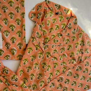 May include: Peach-coloured pyjama set featuring a floral pattern in yellow and green. The set includes a button-down shirt with white piping and matching trousers. The fabric appears soft and comfortable, suitable for sleepwear.