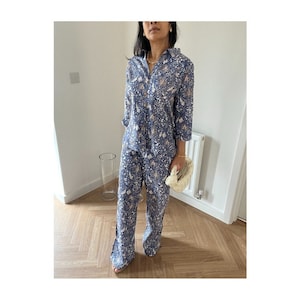 May include: A blue floral patterned pajama set. The set comprises a button-down shirt with three-quarter sleeves and matching wide-leg trousers. A small, cream-colored handbag completes the outfit. The outfit is worn indoors.