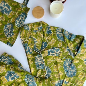 May include: A green floral pajama set with a matching tote bag, a wooden lid, and a white candle. The pajamas have a button-down top with white trim and a pocket. The floral pattern includes blue and yellow accents.