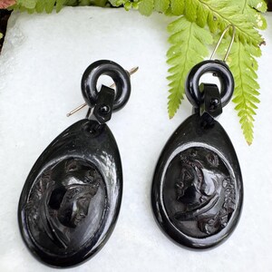 May include: A pair of black jet earrings with a carved cameo design. The earrings are teardrop shaped and have a black metal hook.