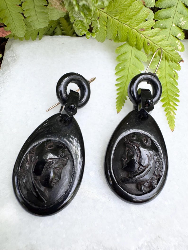 May include: A pair of black jet earrings with a carved cameo design. The earrings are teardrop shaped and have a black metal hook.