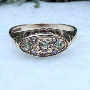 Puede incluir: Anillo dorado con una cara ovalada. La cara presenta tres piedras preciosas verde claro y piedras claras más pequeñas, engastadas en un diseño de estrella. La banda tiene recortes decorativos.