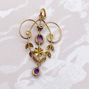 May include: A gold pendant with a delicate, ornate design featuring a central purple stone and smaller purple stones on the sides. The pendant is suspended from a loop at the top.