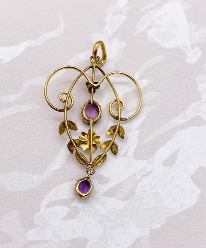 May include: A gold pendant with a delicate, ornate design featuring a central purple stone and smaller purple stones on the sides. The pendant is suspended from a loop at the top.