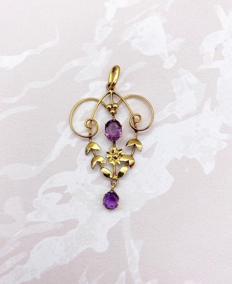 May include: A gold pendant with a delicate floral design and three amethyst stones. The pendant is made of gold and features a floral motif with leaves and a flower. The amethyst stones are oval-shaped and are set in the center of the pendant.
