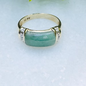 Op de afbeelding: Een gouden ring met een rechthoekige, lichtgroene jade steen. De ring heeft kleine, heldere stenen aan weerszijden van de jade. De ring ligt op een wit oppervlak.