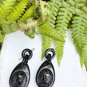 May include: A pair of black jet earrings with a cameo design. The earrings are teardrop shaped and have a gold-tone hook closure.