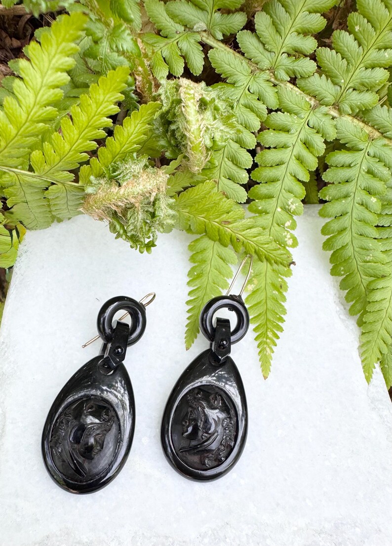 May include: A pair of black jet earrings with a cameo design. The earrings are teardrop shaped and have a gold-tone hook closure.