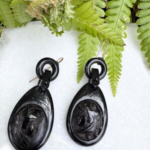 May include: A pair of black jet earrings with a cameo design. The earrings are oval shaped and have a black metal loop at the top.