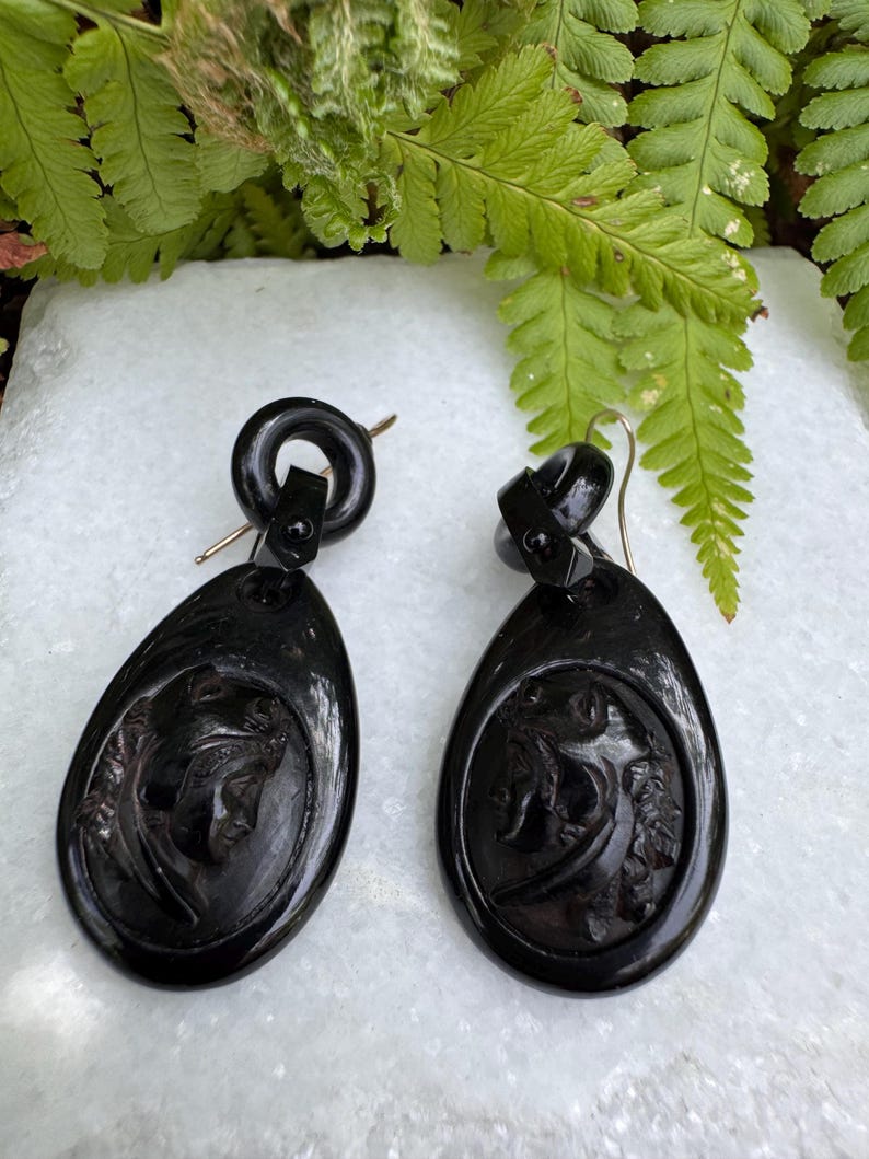 May include: A pair of black carved earrings with a detailed design of a woman's face. The earrings are oval shaped and have a black metal hook.