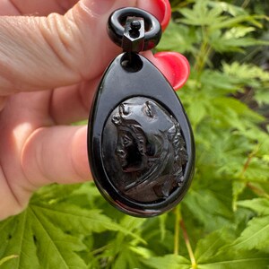 May include: A black cameo pendant with a carved profile of a woman's face. The pendant is attached to a black metal bail.