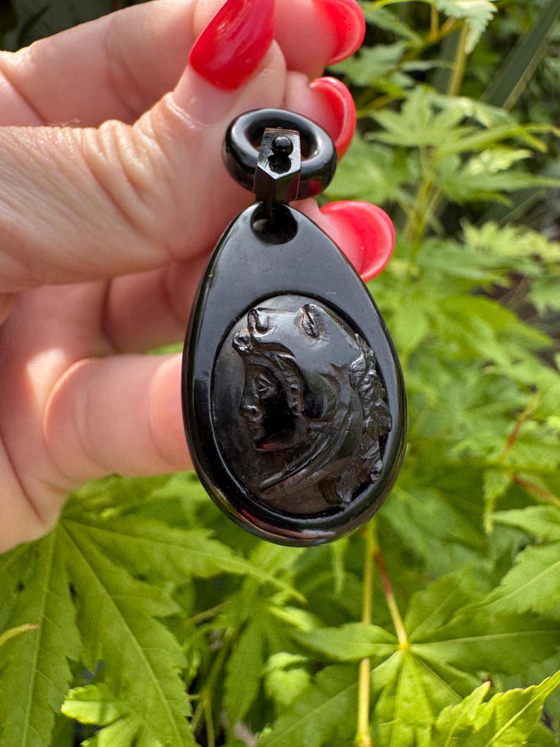 May include: A black cameo pendant with a carved profile of a woman's face. The pendant is attached to a black metal bail.