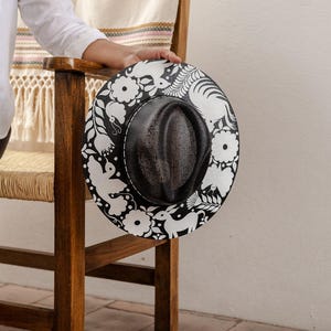 May include: A black and white straw hat with a floral and animal design. The hat has a black mesh crown and a wide brim.