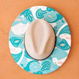 May include: A beige fedora hat with a patterned brim. The brim has a white background with teal and light grey floral and bird designs. The hat has a black band where the brim meets the crown.