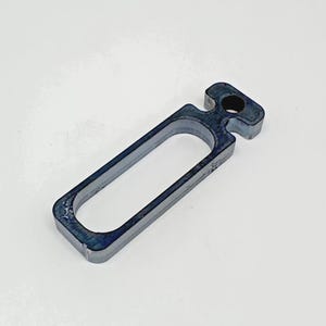 Puede incluir: Un objeto rectangular de plástico azul oscuro con un lazo y un agujero. El objeto tiene una abertura rectangular en el medio. El artículo es probablemente una herramienta o un componente de un producto más grande.