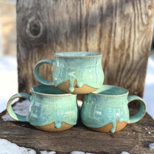 May include: Three handmade ceramic mugs with a turquoise glaze and a natural clay base. The mugs have a rounded shape with a handle and a dripped glaze effect. They are stacked on a weathered wooden surface.