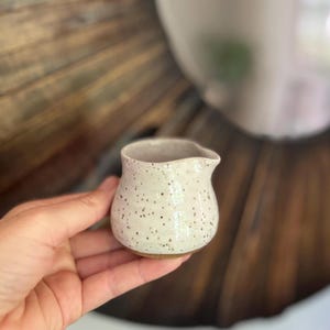 May include: A small, speckled ceramic creamer with a spout, held in a hand. The creamer is off-white with dark brown speckles and a light brown base. The background is a blurred wooden surface.