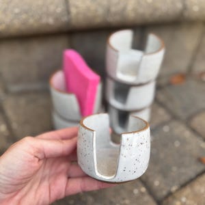 May include: A set of three white ceramic dish soap dispensers with a speckled finish. The dispensers have a unique design with a cut-out for a sponge.