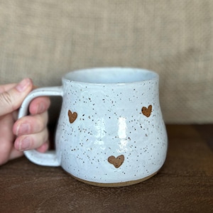 May include: White ceramic mug with a speckled finish and three brown heart-shaped designs. The mug has a rounded shape, a large handle, and a brown base. The mug is held by a hand.