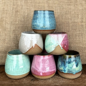 May include: Six ceramic mugs in various colors, including blue, white, pink, and green. The mugs are stacked in a pyramid shape on a brown burlap background.