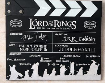 May include: A black and white film clapperboard for "The Lord of the Rings: The Fellowship of the Ring." Features the film's title, director Peter Jackson, and the cast, including Elijah Wood. The clapperboard has a black body with white stripes.